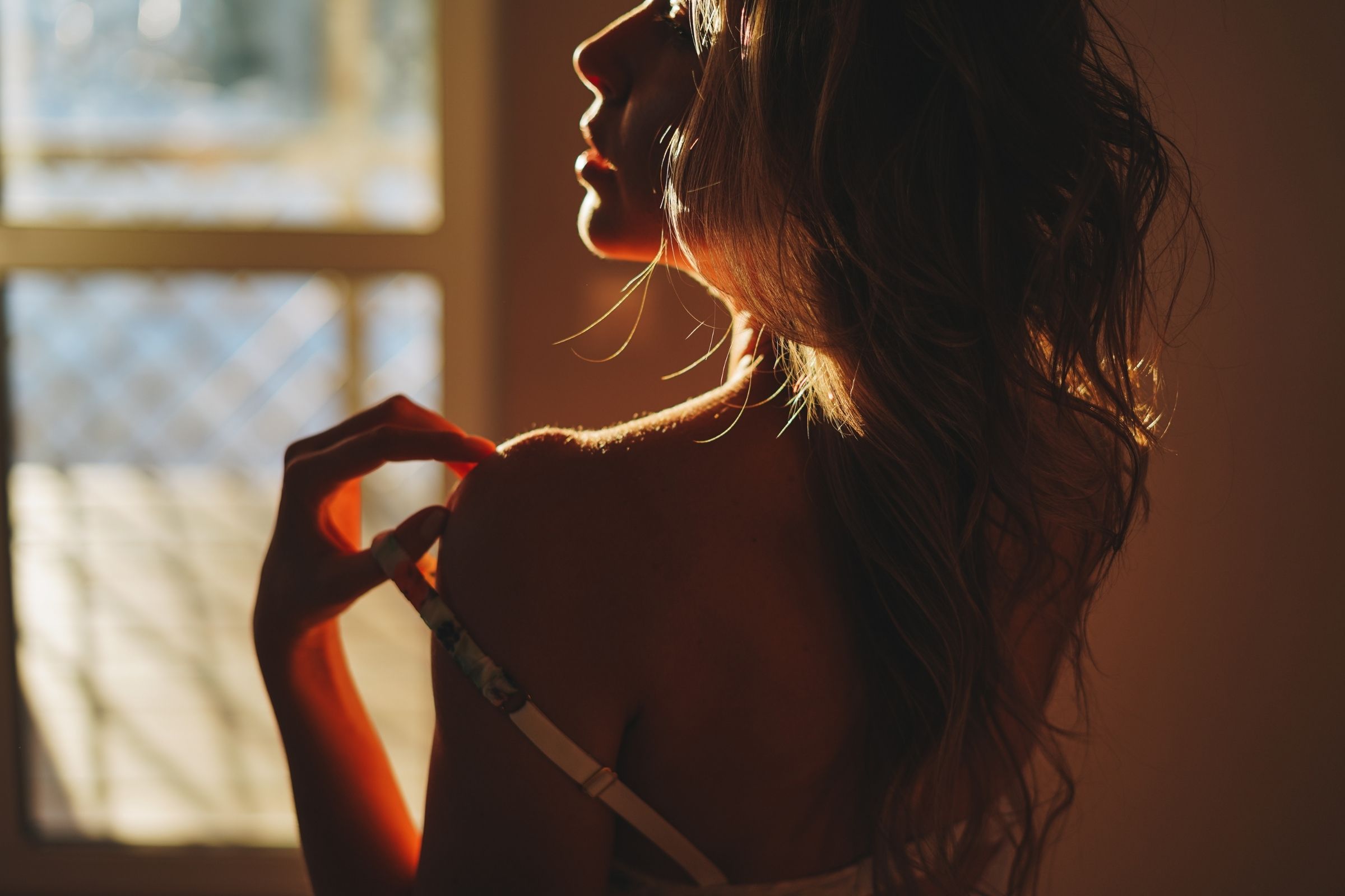 Woman in profile touching her shoulder in amber light beside a window